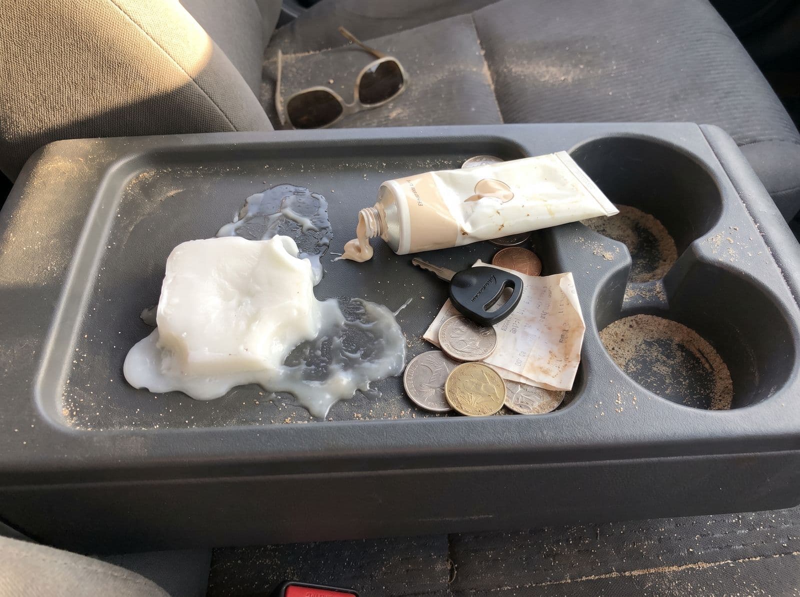 A messy car centre console: half-melted surf wax stuck to the plastic, a squeezed zinc tube, keys, sandy coins and a crumpled receipt